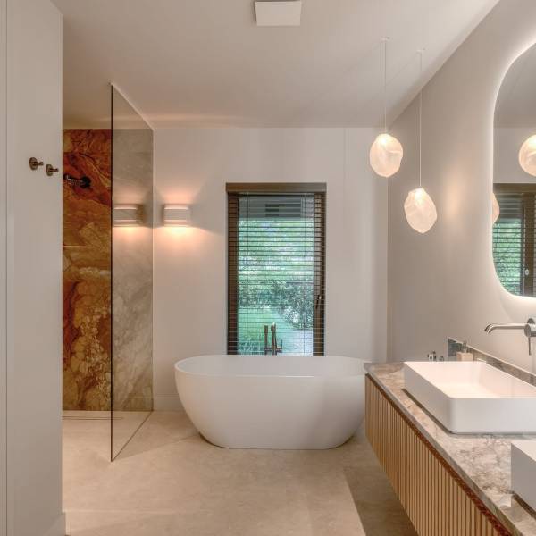 Badkamer met vrijstaand wit bad, warmgekleurde douchewand, geribbeld wastafelmeubel en hanglampen.