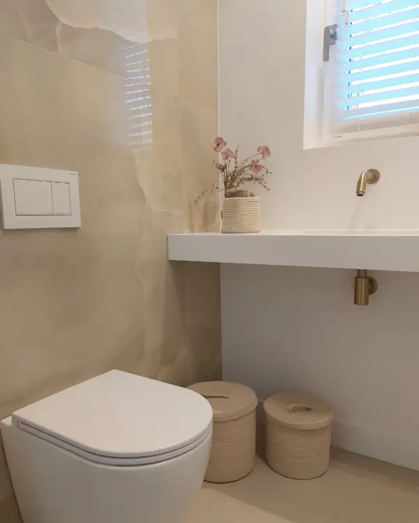 Toiletruimte met grote glanzende beige wandtegels, wit sanitair, een goudkleurige wandkraan en gevlochten manden.