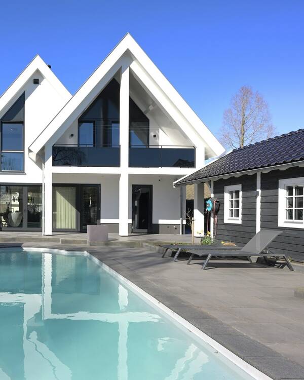 Grijze terrastegels rond een zwembad bij een witte woning met donkere kozijnen, een zwart bijgebouw en ligstoelen.