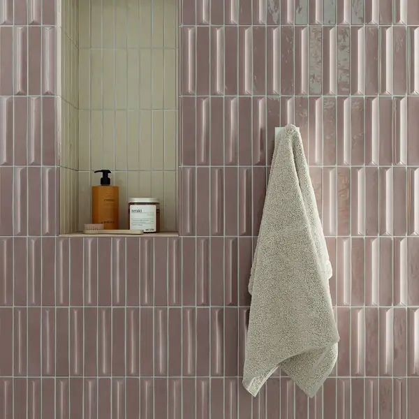 Zachtroze wandtegels met reliëf, een licht betegelde nis met verzorgingsproducten en een beige handdoek.