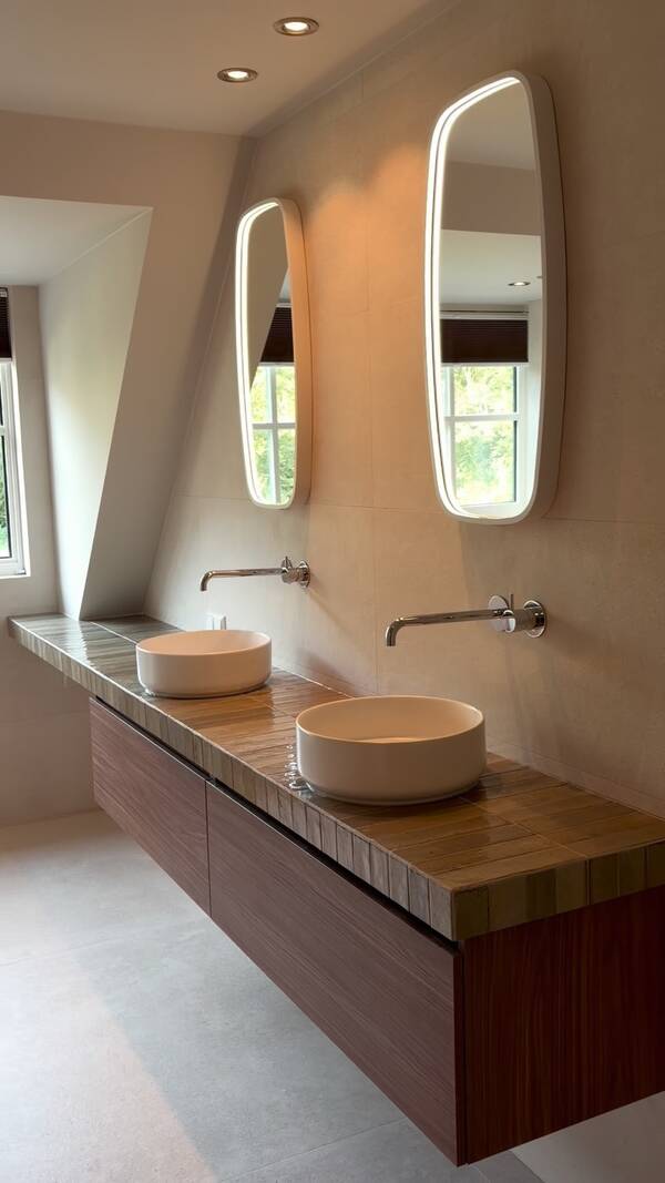 Badkamer met betegeld wastafelblad, twee ronde waskommen, meubel met houtlook en langwerpige verlichte spiegels.